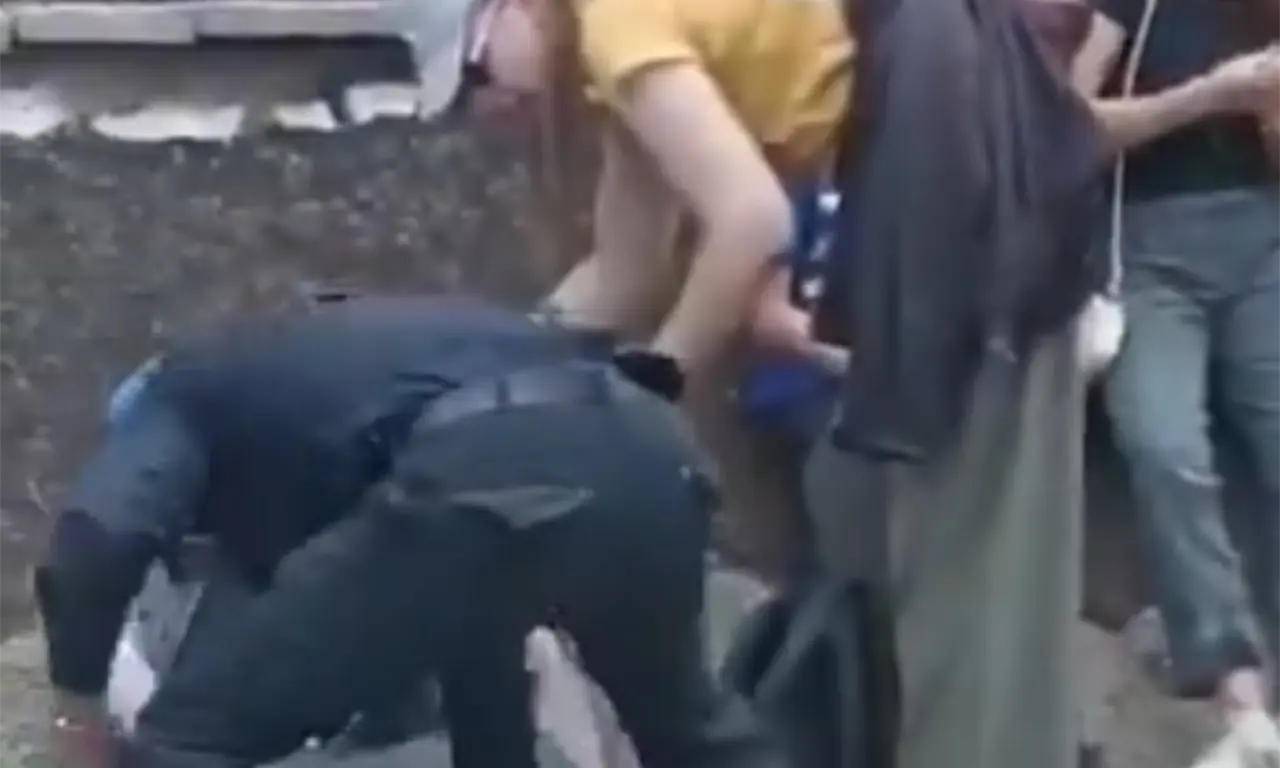  En el centro de la escena, un hombre con la pierna ensangrentada permanece recostado mientras quienes lo rodean buscan mantenerlo consciente y con vida.
 